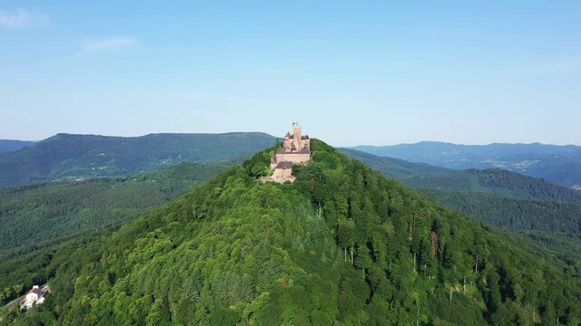 Drone shot of Chateau du Haut Koenigsbourg atop a lush green hill surrounded by forested mountains in Alsace, France.
