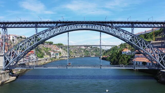 Wide view of the iconic Dom Lu&iacute;s I Bridge spanning the Douro River in Porto, Portugal, with colorful riverside buildings and lush hillsides under a clear blue sky.