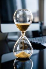 Glass sand timer counting down minutes on busy office desk with computer