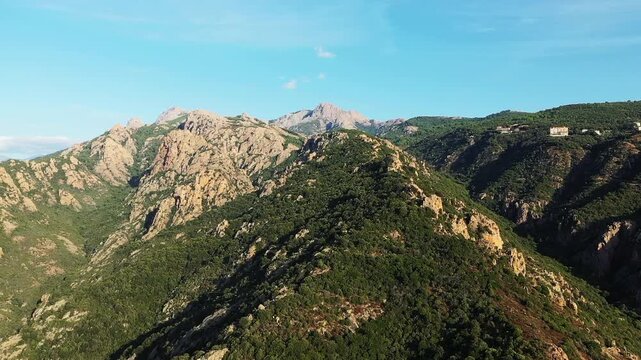 Aerial view of rugged green mountains and rocky peaks in the Corsican countryside under a clear blue sky. Scenic natural terrain with lush vegetation and dramatic elevation.