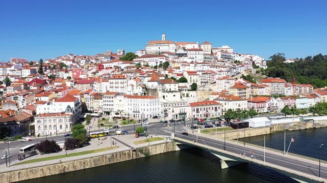 Scenic view of Coimbra with historic buildings and the Mondego River under a clear blue sky. The city&rsquo;s hillside architecture and riverside bridge create a picturesque urban landscape.