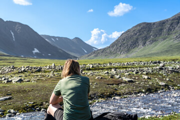 Woman enjoys the view from a rocky riverbank in Trollheimen. High mountains rise in the distance under a blue sky with a few clouds. Hiking in Norway
