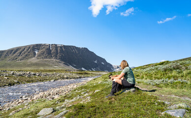 A female hiker sits by a river in the Trollheimen mountain area of Norway. Bl&aring;h&oslash;a mountain is in the background. Snow is visible in the summer landscape.
