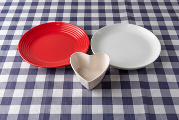 Red plate, white plate and a heart-shaped bowl on a blue and white checkered tablecloth. Romantic dining and colorful tableware concept