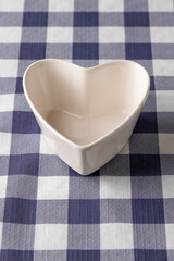 Empty white heart-shaped ceramic bowl on a blue and white checkered tablecloth. Romantic tableware for snacks, desserts or Valentines Day