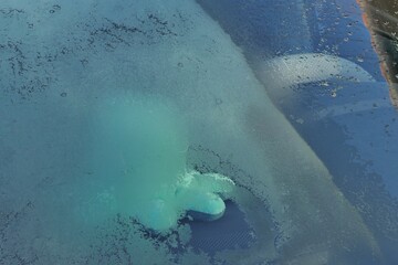 Half frozen and half melted front view of modern C segment sedan car during wintertime. There is driving wheel with beige cover and cyan coloured plush octopuss visible behid the window