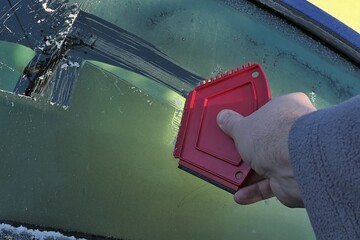 Hand of adult male person scratching down frost cover from side window of modern C-segment car with multi purpose compact red plastic scraper. 