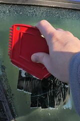 Hand of adult man scratching down frost cover from side window of modern C-segment sedan car with multi purpose compact red plastic scraper. 