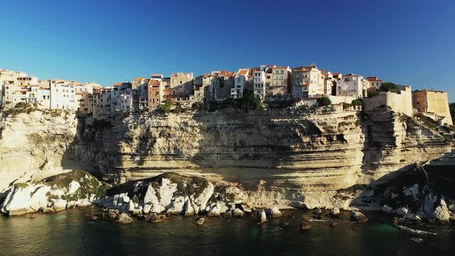 Dramatic view of colorful houses perched atop limestone cliffs overlooking the sea in Bonifacio, Corsica, under a clear blue sky.
