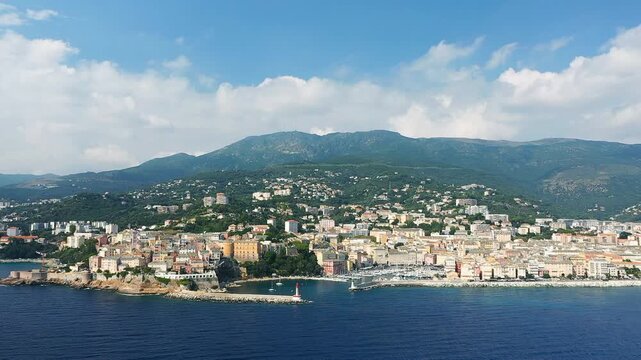 Scenic aerial view of Bastia with colorful buildings along the coastline and lush green mountains in the background, under a bright blue sky.