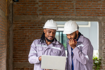 Two Black construction workers in safety helmets review plans on a laptop at a brick building site, highlighting teamwork, technology and workplace diversity in the construction industry. Team work