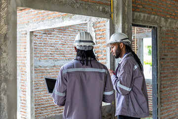 Two Black construction workers in safety helmets review plans on a laptop at a brick building site, highlighting teamwork, technology and workplace diversity in the construction industry. Team work