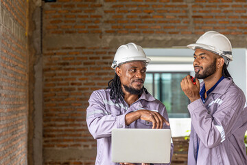 Two Black construction workers in safety helmets review plans on a laptop at a brick building site, highlighting teamwork, technology and workplace diversity in the construction industry. Team work