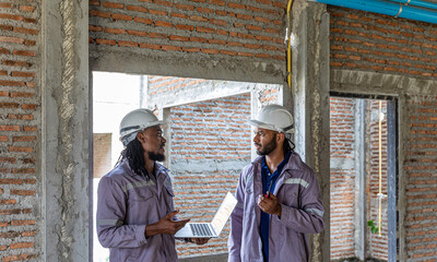 Two Black construction workers in safety helmets review plans on a laptop at a brick building site, highlighting teamwork, technology and workplace diversity in the construction industry. Team work