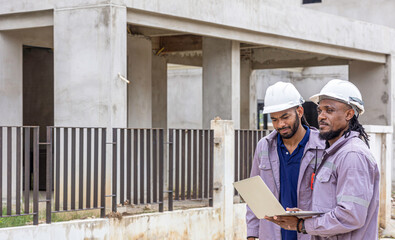 Two Black construction workers in safety helmets review plans on a laptop at a brick building site, highlighting teamwork, technology and workplace diversity in the construction industry. Team work