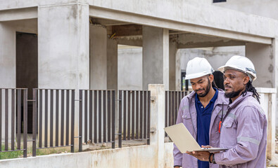 Two Black construction workers in safety helmets review plans on a laptop at a brick building site, highlighting teamwork, technology and workplace diversity in the construction industry. Team work