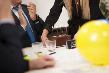 Office birthday celebration with chocolate cake, lit candles, yellow balloons, and colleagues clapping as friends celebrate a work anniversary at the desk in a friendly, professional setting