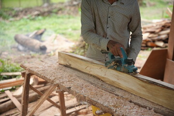 A carpenter shaping a wooden plank using an electric planer, working outdoors as wood shavings scatter, showing a hands on woodworking process in a natural workshop setting.