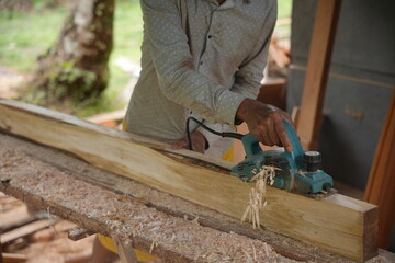 A carpenter shaping a wooden plank using an electric planer, working outdoors as wood shavings scatter, showing a hands on woodworking process in a natural workshop setting.