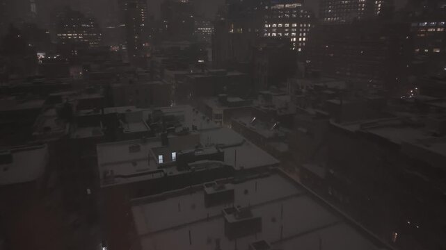 A cinematic aerial drone shot glides forward over snow-covered rooftops in New York City during a heavy night blizzard. Warm glowing lights from windows and street lamps contrast against the cool, ...