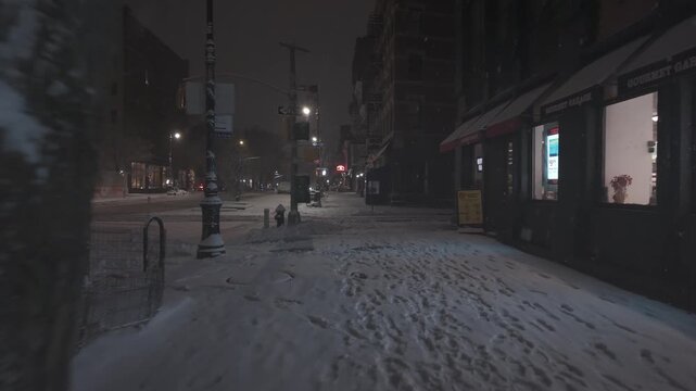 Low-altitude drone footage glides down a snow-covered Manhattan sidewalk during a quiet night storm. The cinematic dolly-in movement captures the warm glow of storefront windows contrasting against...