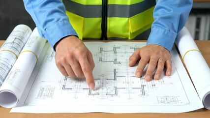 Hands of an engineer in a safety vest pointing at an architectural blueprint on a desk with rolled drawings, concept for construction planning, design review and industrial development - Powered by Adobe