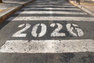 The year 2026 is prominently stenciled in bold white paint on a textured asphalt road, forming part of a pedestrian crosswalk. The numerals stand out against the dark pavement, displaying a slightly w