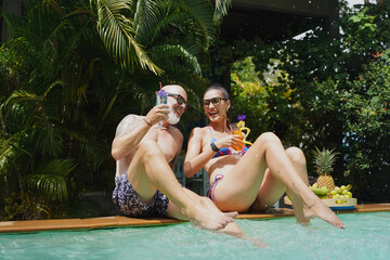 A man and a woman sit by the pool with cocktails and fruit.