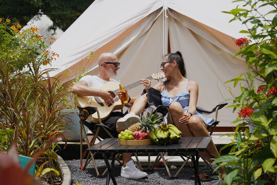 A man plays guitar for a woman near a cozy tent at a glamping site.
