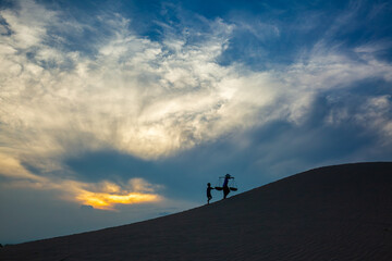 Cham women walking home across the sand dunes at sunset, beneath a blue sky filled with soft white clouds.