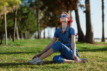 Fototapeta premium young woman sitting on grass outdoors wearing blue denim jumpsuit and sunglasses enjoying warm sunny day in green park environment