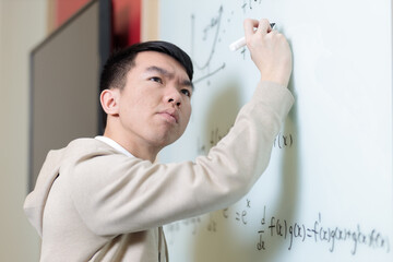 A Chinese student concentrates while writing mathematical formulas on a whiteboard with a marker. He is working through complex equations in a classroom.