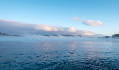 Serene lake mist rises, soft clouds drift above calm waters under clear blue sky.