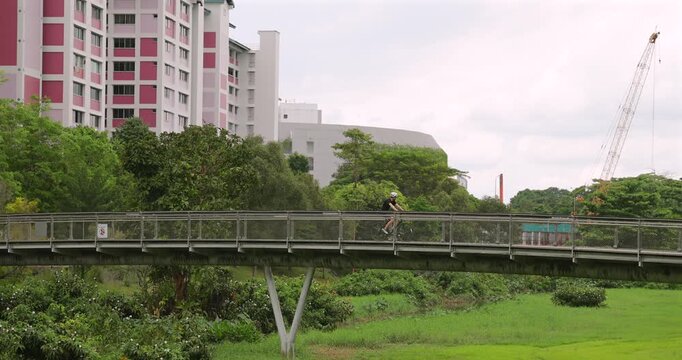 Man cycles across elevated pedestrian bridge in lush urban park. Modern residential buildings, construction crane, overcast sky. Healthy lifestyle, city infrastructure.