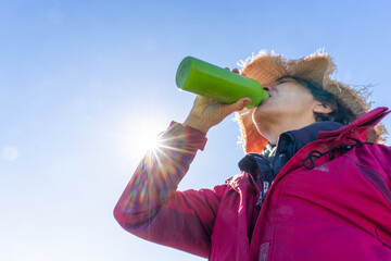 Person wearing a hat and jacket drinking from a green reusable water bottle in bright sunlight,...