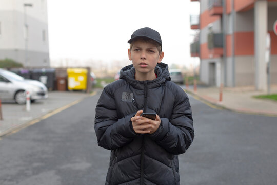 young white male checking phone outdoors near residential block, cap on, puffer jacket, focused expression, urban sidewalk, muted overcast light, casual city routine vibe - Powered by Adobe