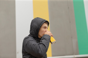 Profile caucasian young man drinking from can, hood up over dark puffer jacket, standing by yellow stripe mural, quick urban break, focused gaze toward street, candid refreshment moment,