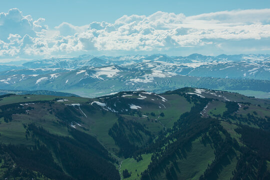 Aerial sunlit top view above forest hills and big snowy mountain range under lush clouds in changeable weather. Scenic hilly alpine landscape with sunlight and shadows. Partly cloudy in high mountains