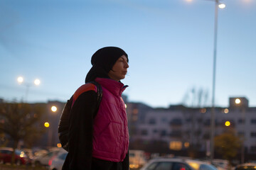 young adult profile framed by night lights wearing bright vest and backpack, cool city evening with illuminated streetlamps and distant building silhouettes, thoughtful nighttime mood