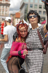 two women posing smiling for camera in busy festival square, red hair accent, costume details, warm laughter,