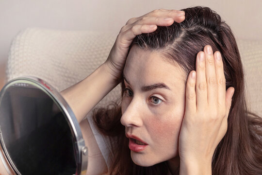 Young brunette woman, looks in the mirror at the first gray hair on her head. She's in shock. Copy space photo