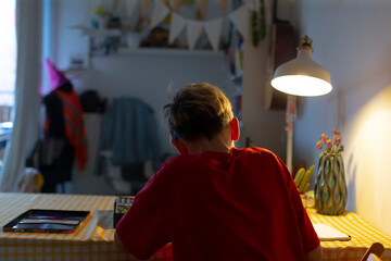 Teenage boy studying at desk under lamp, wearing red sweatshirt and headphones while writing in notebook on checkered table, warm lamp glow casts soft shadows, tidy bedroom