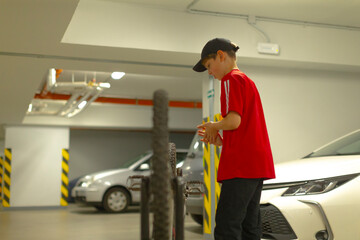 teen carrying spare bicycle wheel through garage toward workshop, dressed in red shirt and cap while stepping past parked car with determined posture motion and grip emphasize action,