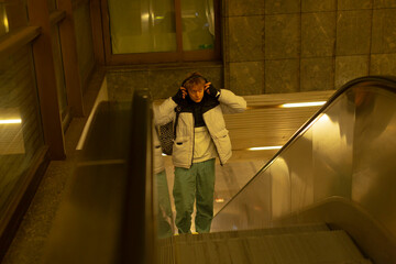 White man adjusting headphones on escalator, playful candid moment with hands near head, puffer jacket and tote bag, upward movement under warm artificial light, thoughtful expression