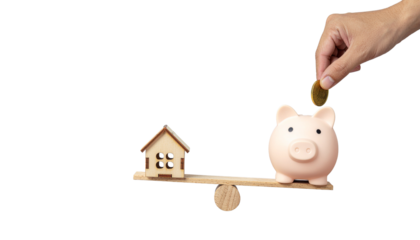 Hand places coin into piggy bank on seesaw opposite small house, illustrating financial balance no background
