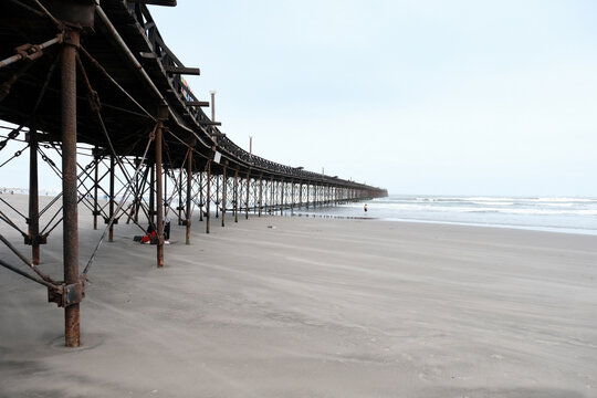 Rusty iron structure of pimentel pier stretching across the vast sandy beach under an overcast sky with ocean waves