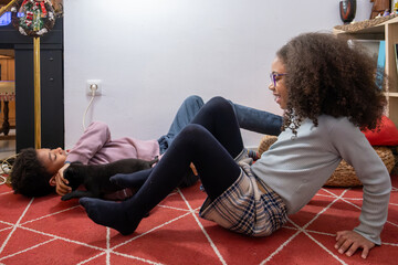 Two mixed-race children lounging on a carpet, joyfully playing and bonding with a small black kitten in a cozy home setting