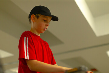 young boy wearing red shirt adjusting training equipment indoors, wearing black cap, focused expression under warm fluorescent lights, candid portrait showing preparation, concentration