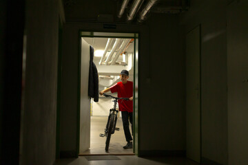 messenger at doorway holding bicycle, redjacketed courier pauses at threshold between corridor and street, sunlight contrasts interior shadows creating dynamic urban delivery moment