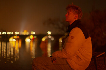young man seated by river under amber lights, profile in casual jacket with shimmering water reflections and soft bridge bokeh, contemplative pose and calm expression, cinematic warm tones,